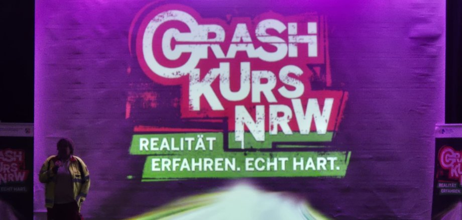 Снимка на сцената с голямо лого "Crash Kurs NRW" и слоган "Изживейте реалността. Наистина трудна."; вляво е служител на спешна помощ в облекло с висока видимост, отдолу се виждат зрители.