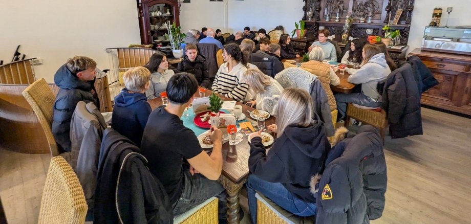 В голяма зала на Café Grenzenlos ученици от 9 и 10 клас седят заедно с по-възрастни участници в инициативата "Бабите срещу десницата". Хората са разпределени в стаята на няколко маси и седят един срещу друг в малки дискусионни групи. Някои седят на столове до маси с напитки и чинии, други - на дивани покрай стената. На заден план се виждат други групи, които също разговарят помежду си.