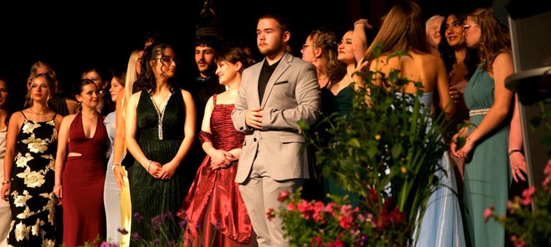 Na pódiu stojí slavnostně oblečená skupina mladých lidí a dívá se stranou, jako by čekala na další oznámení programu. Lidé mají na sobě elegantní šaty a obleky, v popředí jsou vidět velké květináče s barevnými květinami. Jeviště je tmavě osvětleno a skupina je osvětlena teplými reflektory, což vytváří slavnostní atmosféru.