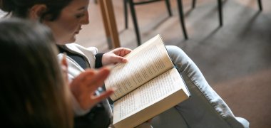 V synagoze sedí na židli žákyně ze základní školy v Gronau, v rukou drží otevřenou knihu s hebrejským písmem a čte si v ní, zatímco vedle ní jiná osoba gestikuluje a něco vysvětluje.