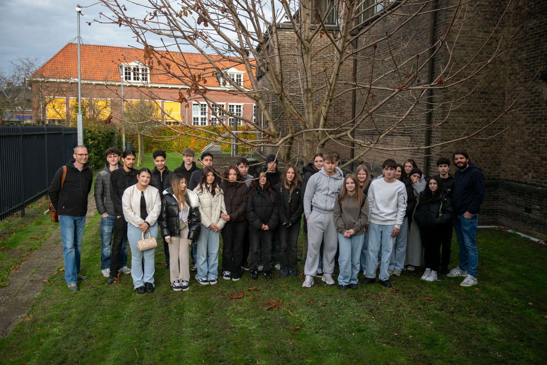 En stor gruppe elever fra 9.-10. klasse på Gronau grundskole står sammen med to lærere på plænen ved siden af synagogen i Enschede; bag dem ses et nøgent træ, synagogens murstensvæg og beboelsesejendomme med røde tage.