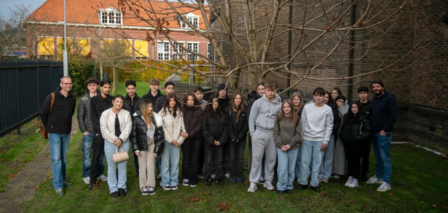 Besøg i synagogen i Enschede - 2025 En stor gruppe elever fra 9.-10. klasse på Gronau grundskole står sammen med to lærere på plænen ved siden af synagogen i Enschede; bag dem ses et nøgent træ, synagogens murstensvæg og beboelsesejendomme med røde tage.