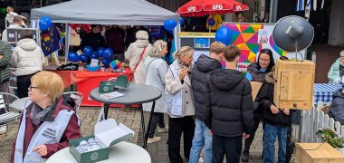 På en plads står flere mennesker ved forskellige boder på en gadefest. I forgrunden står et rundt højbord med flyers, badges og en åben kasse. Til venstre står en person iført en lys vest fra initiativet »Omas gegen Rechts«. I midten af billedet er flere unge og voksne i gang med at tale sammen. Bag dem ses et telt med blå balloner, informations- og aktivitetsstande samt en fotoboks. I baggrunden ses butikker, siddepladser og flere besøgende på festivalen. Scenen giver indtryk af en velbesøgt og åben udveksling mellem forskellige generationer.