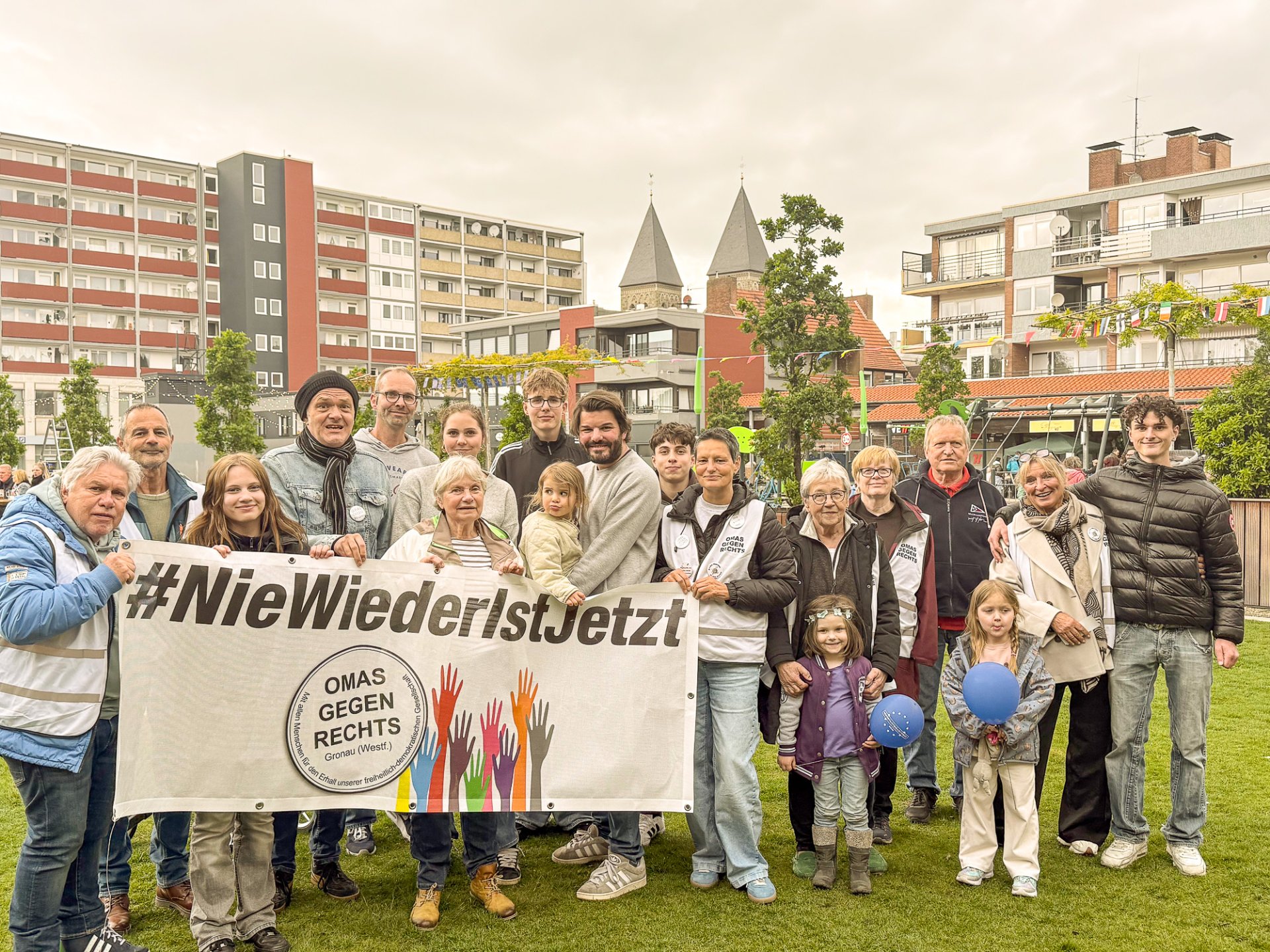 På en græsplæne står omkring tyve mennesker i forskellige aldre samlet til et gruppebillede. I forgrunden holder flere personer et stort banner med påskriften »#NieWiederIstJetzt« samt logoet for Gronau-afdelingen af »Omas gegen Rechts«. Gruppen består af ældre og yngre voksne, unge samt flere børn. Nogle bærer badges eller veste fra initiativet. I baggrunden ses boligbygninger, træer og tårnene på en kirke. Billedet blev taget i forbindelse med Europafesten på Platz der Kulturen i Gronau.