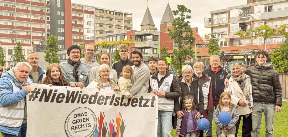 På en græsplæne står omkring tyve mennesker i forskellige aldre samlet til et gruppebillede. I forgrunden holder flere personer et stort banner med påskriften »#NieWiederIstJetzt« samt logoet for Gronau-afdelingen af »Omas gegen Rechts«. Gruppen består af ældre og yngre voksne, unge samt flere børn. Nogle bærer badges eller veste fra initiativet. I baggrunden ses boligbygninger, træer og tårnene på en kirke. Billedet blev taget i forbindelse med Europafesten på Platz der Kulturen i Gronau.