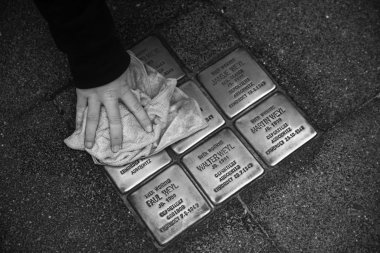 Close-up black and white photo: A pupil's hand from Gronau Comprehensive School wipes a cloth over several Stumbling Stones on November 4, 2025, so that the names and dates of the Jewish victims can be clearly recognized as part of the Week of Remembrance.