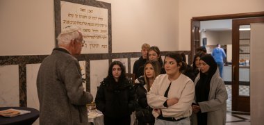 Inside the synagogue: an older volunteer with a kippah speaks to a group of Year 9/10 pupils from Gronau comprehensive school, who listen attentively; a large Hebrew inscription hangs on the wall in the background.