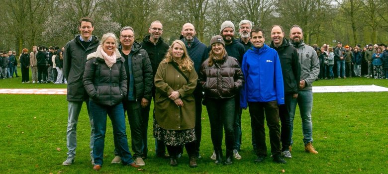 A group of twelve adults stand on a meadow in the foreground and look into the camera. They are teachers and representatives of various schools and institutions. Behind them, a large human chain of many pupils stretches across the school grounds. The people are standing close together on a green area, some wearing jackets and coats. Trees with fresh leaves and an overcast sky can be seen in the background. The scene shows a collective action on the school grounds.