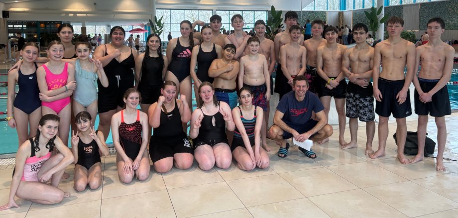 A large group of children and young people in swimwear stand and kneel together at the edge of a modern indoor swimming pool. The group smiles and poses for a photo; some show hand signs. The pool, large window fronts with a view outside and other bathers can be seen in the background. An adult trainer kneels to the front right of the group.