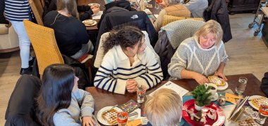En una sala del Café Grenzenlos, los alumnos se sientan en una gran mesa junto a participantes de más edad en la iniciativa "Abuelas contra la derecha". Sobre la mesa hay platos de gofres, vasos y tazas. Varios jóvenes se sientan en sillas alrededor de la mesa, mientras que los participantes de más edad se sientan a su lado y también comen o hablan. Al fondo hay un gran armario de madera oscura con figuras talladas y adornos. Una persona entrega una taza a un participante sentado. Se pueden ver otros grupos de discusión en una segunda mesa de la sala.