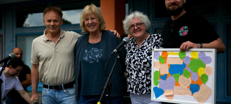 schools-out-04 Neli inimest seisavad käsivarrelt laval koolimaja ees ja naeratavad kaamerasse. Vasakult paremale: Härra Schnigge, proua Steuer (direktor), proua Holtkamp ja härra Gloddek. Härra Gloddek hoiab käes valget pildiraami värviliste hüvastijätusõnumitega.