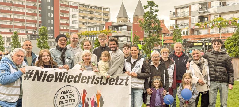 Muruplatsil seisab umbes kakskümmend erinevas vanuses inimest grupifoto tegemiseks koos. Esiplaanil hoiavad mitu inimest suurt bännerit, millel on kiri „#NieWiederIstJetzt“ ning Gronau kohaliku rühmituse „Omas gegen Rechts“ logo. Rühma kuuluvad vanemad ja nooremad täiskasvanud, noored ning mitu last. Mõned neist kannavad algatuse märgikesi või veste. Taustal on näha elumaju, puid ja kiriku torne. Foto on tehtud Euroopa festivali raames Gronau Kultuuride väljakul.