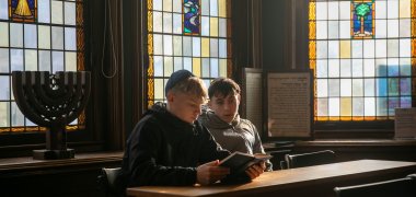 Visite de la synagogue d'Enschede - 2025 Deux élèves de 9e/10e année, kippa sur la tête, sont assis à une table en bois dans la synagogue d'Enschede et feuillettent un livre de prières. Un chandelier de la Menorah est posé à côté d'eux et de grands vitraux colorés brillent derrière eux.