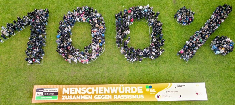 Une vue à vol d'oiseau montre un grand groupe d'élèves* qui se tiennent proches les uns des autres et forment le chiffre "100 %". Les chiffres et le signe du pourcentage sont clairement identifiables grâce à la disposition des personnes. En dessous de la formation se trouve une longue bannière sur laquelle on peut lire "Dignité humaine - ensemble contre le racisme". La photo montre l'ensemble du groupe vu d'en haut et fait apparaître clairement la forme du chiffre.