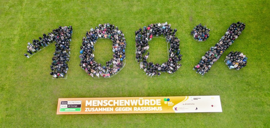 Une vue à vol d'oiseau montre un grand groupe d'élèves* qui se tiennent proches les uns des autres et forment le chiffre "100 %". Les chiffres et le signe du pourcentage sont clairement identifiables grâce à la disposition des personnes. En dessous de la formation se trouve une longue bannière sur laquelle on peut lire "Dignité humaine - ensemble contre le racisme". La photo montre l'ensemble du groupe vu d'en haut et fait apparaître clairement la forme du chiffre.