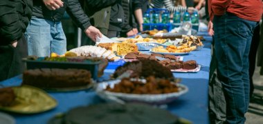 Une longue table recouverte d'une nappe bleue est garnie de divers gâteaux, pâtisseries et autres mets. Plusieurs élèves se tiennent de part et d'autre du buffet. Certains se servent, d'autres examinent ce qui est proposé. À l'arrière-plan, on aperçoit d'autres personnes ainsi que des caisses de boissons.