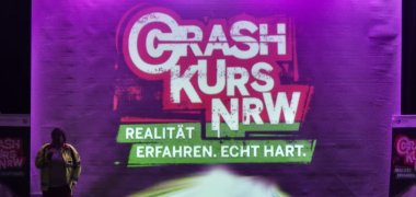 Photo de scène avec un grand logo "Crash Kurs NRW" et le slogan "Expérimenter la réalité. Vraiment dur" ; à gauche, un agent d'intervention en tenue d'avertissement ; en bas, des spectateurs*.