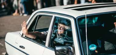 Tiba dengan mobil klasik Ursula Steuer duduk sambil tersenyum di bangku belakang sebuah mobil klasik berwarna putih dan melambaikan tangan dari jendela samping yang terbuka. Di latar belakang, para siswa, guru, dan staf tampak samar-samar berdiri di halaman sekolah sambil menanti kedatangannya.