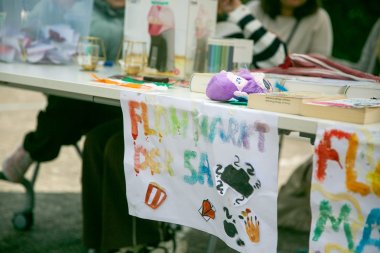 Di atas sebuah meja terdapat berbagai barang, antara lain buku, boneka, dan barang-barang pasar loak lainnya. Di bagian depan meja tergantung sebuah poster buatan sendiri bertuliskan “Pasar Loak”. Di latar belakang, beberapa siswa dan siswi duduk dan berdiri di belakang stan tersebut.
