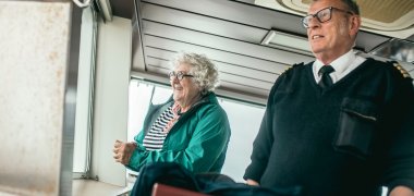Di ruang kemudi kapal feri, kapten dan seorang guru dari Sekolah Menengah Gronau berdiri berdampingan di dekat jendela. Keduanya tersenyum dan memandangi laut selama perjalanan pulang. Kapten mengenakan seragamnya, sementara di latar depan terlihat sebagian ruang kemudi.