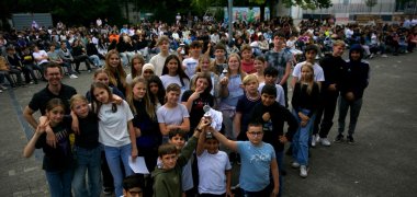 Un gruppo numeroso di alunni si trova vicino in un'area di gioco della scuola, alcuni con certificati o trofei in mano. In primo piano, diversi bambini mostrano un trofeo alla telecamera. Un pubblico numeroso siede su delle sedie sullo sfondo.