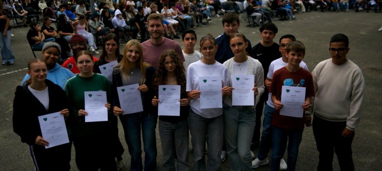 Gli alunni dell'Istituto Comprensivo di Gronau sono in piedi in due file nel cortile della scuola. Tengono i certificati in mano e sorridono alla telecamera. Il signor Nöthe, l'insegnante responsabile, è in piedi in mezzo a loro. Sullo sfondo c'è il pubblico nel cortile della scuola.