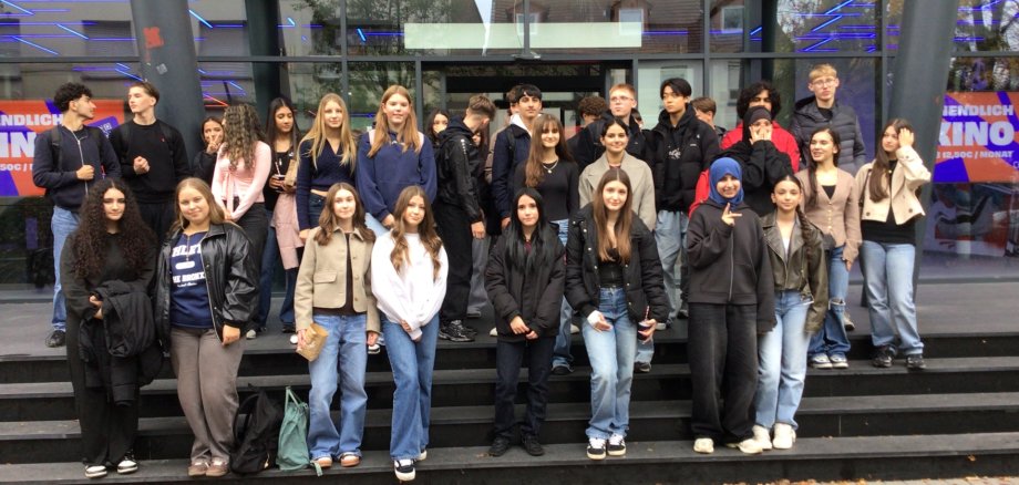 Un folto gruppo di alunni dell'Istituto Comprensivo di Gronau e dello Stedelijk Lyceum Enschede Zuid si trova insieme sui gradini di fronte al cinema Cinetech di Gronau. I giovani posano per una foto di gruppo dopo una discussione comune sul tema della sostenibilità. La facciata in vetro del cinema con la scritta illuminata "Cinetech" è visibile sullo sfondo.