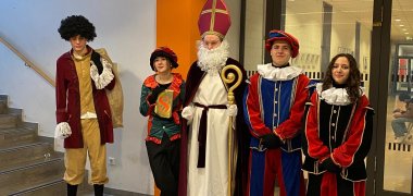 Una persona vestita da Sinterklaas con mitra e pastorale si trova nel corridoio dell'Istituto Comprensivo di Gronau con quattro alunni vestiti da compagni tradizionali. Il gruppo indossa costumi colorati e si prepara alla visita delle classi di quinta elementare.