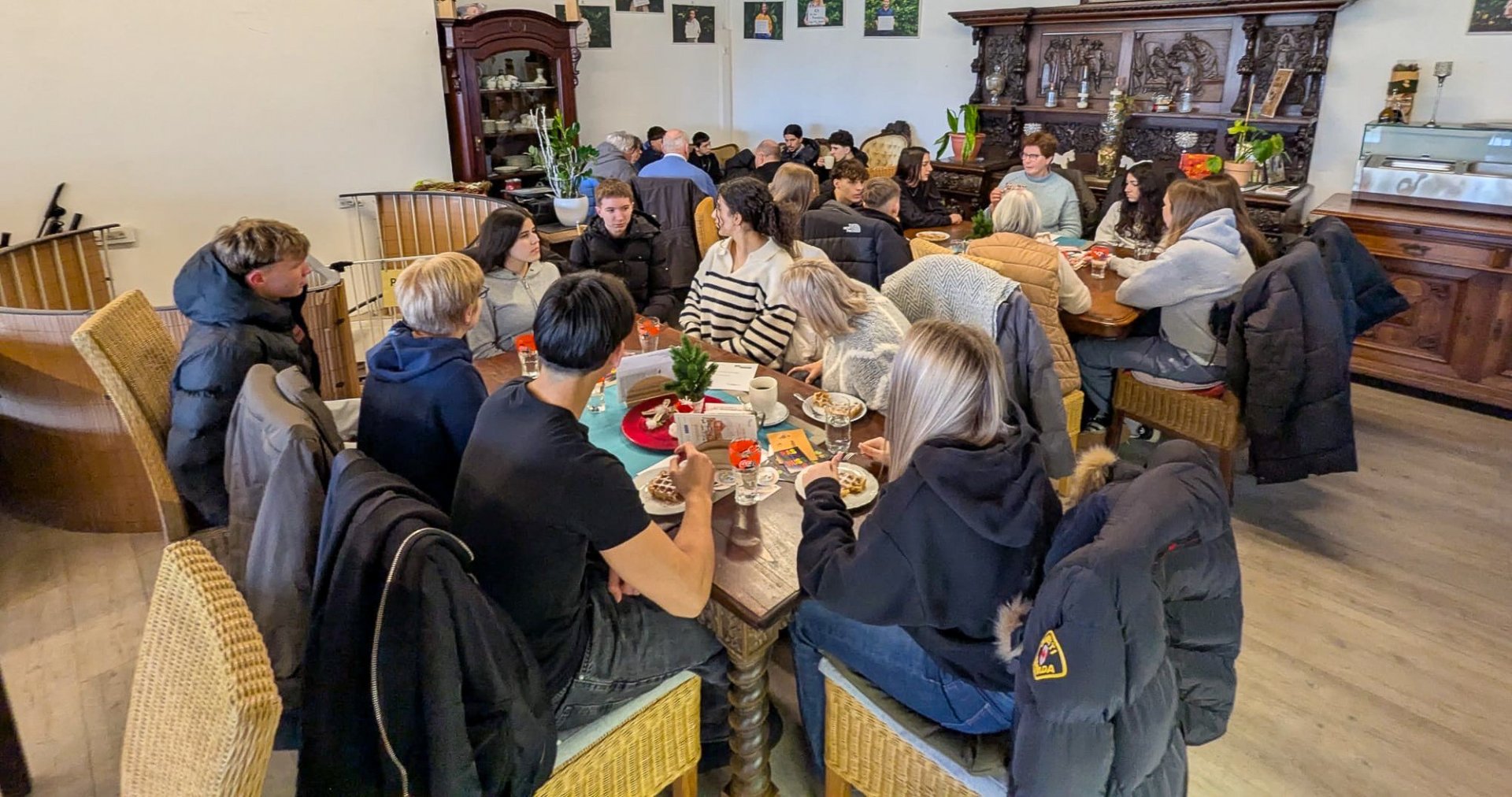 In una grande sala del Café Grenzenlos, gli alunni degli anni 9 e 10 siedono insieme ai partecipanti più anziani dell'iniziativa "Nonne contro la destra". Le persone sono sparse per la stanza in diversi tavoli e siedono l'una di fronte all'altra in piccoli gruppi di discussione. Alcuni sono seduti su sedie a tavoli con bevande e piatti, altri su divani lungo la parete. Sullo sfondo si vedono altri gruppi, che parlano tra loro.