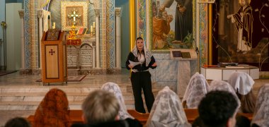 Gausiai išdažytoje sirų ortodoksų bažnyčioje Gronau mieste mokinė Hala stovi priešais altorių su baltu nėriniuotu šydu ir kalbasi su suoluose sėdinčiais 9-10 klasių mokiniais, kai kurie iš jų taip pat dėvi šydus; fone matyti kryžius, ikonos ir žvakės.