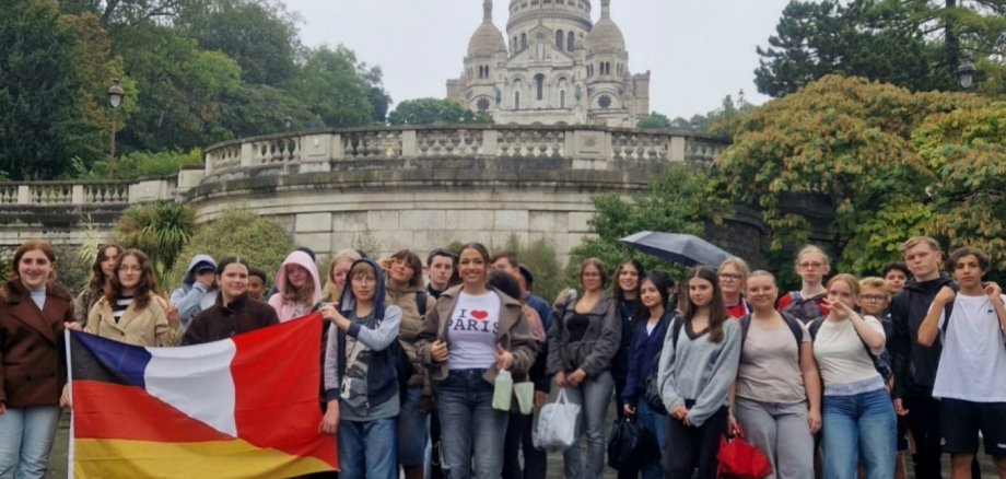 Mokinių grupė, vykstanti į ekskursiją į Prancūziją, priešais Sacré-Cœur baziliką Monmartre kalne Paryžiuje. Kai kurie paaugliai laiko vokiečių-prancūzų vėliavą. Fone matyti šviesi kupolinė bažnyčia ir apželdinta terasa.