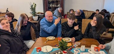 Keletas mokinių sėdi prie didelio medinio stalo kavinėje "Grenzenlos" kartu su vyresniu iniciatyvos "Senelės prieš dešiniuosius" dalyviu. Ant stalo guli vaflių lėkštės, stiklinės, puodeliai ir įvairios skrajutės bei kortelės. Jaunuoliai susėda ant kėdžių aplink stalą ir sutelkia dėmesį į pokalbį. Centre sėdintis vyresnis dalyvis kalba ir gestikuliuoja rankomis, o jaunuoliai klausosi arba šypsosi. Fone kiti dalyviai sėdi ant sofos ir prie kito kambario stalo. Už jų stovi didelė tamsi spinta su stikline spintele.