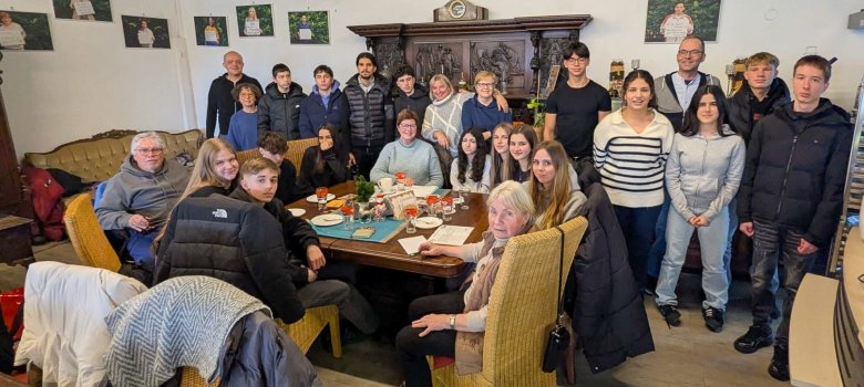 Kafejnīcas "Grenzenlos" telpā skolēni stāv un sēž ap lielu galdu, lai kopā ar vecākiem iniciatīvas "Vecmāmiņas pret tiesībām" dalībniekiem kopīgi nofotografētos. Daži jaunieši sēž uz krēsliem ap galdu, citi stāv aiz galda. Uz galda ir glāzes, tasītes, šķīvji un dažas vafeles. Fonā atrodas liels tumša koka skapis ar grieztiem rotājumiem. Pie sienām karājas vairākas fotogrāfijas, un pa istabu ir izkaisītas citas sēdvietas mēbeles, piemēram, dīvāni un krēsli.