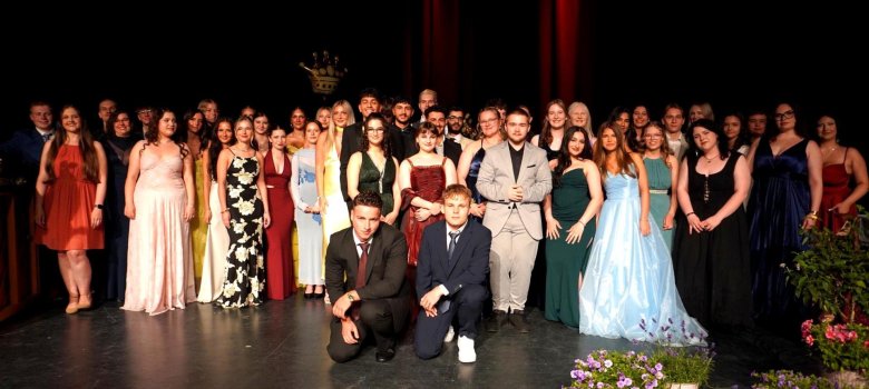 Gruppebilde fra et skoleavslutningsball: Rundt 45 festkledde avgangselever står på en scene foran en mørk bakgrunn og rød scenelys. De unge voksne er iført elegante aftenkjoler, dresser og festlige frisyrer. To studenter i dress og slips kneler i forgrunnen. Stemningen er høytidelig og stolt. Scenekanten er dekorert med blomster, og i bakgrunnen skimtes en gyllen krone.