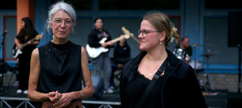Mrs Knowles (assisterende rektor) og Clara Henschke (studentrepresentant) står ved siden av hverandre og smiler inn i kameraet. Et studentband spiller på en liten scene i bakgrunnen.