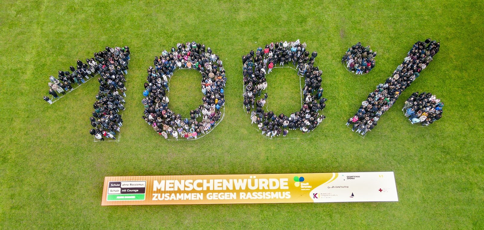 In vogelvluchtperspectief ziet u een grote groep leerlingen die dicht bij elkaar staan en het getal "100 %" vormen. De cijfers en het procentteken zijn duidelijk herkenbaar door de opstelling van de mensen. Onder de formatie hangt een lang spandoek met het opschrift "Menselijke waardigheid - samen tegen racisme". De foto toont de hele groep van bovenaf en maakt de vorm van het getal duidelijk zichtbaar.