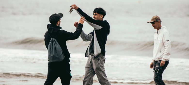Drie scholieren staan op het strand aan de Noordzee. Een van hen houdt een klein krabje omhoog en laat het aan zijn klasgenoten zien, die er nieuwsgierig op reageren. Op de achtergrond zijn de golven en de bewolkte horizon te zien. De foto toont een spontaan moment van ontdekking tijdens het verblijf op Norderney.