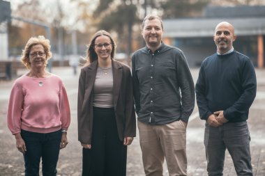 Vier mensen staan naast elkaar in de buitenruimte van een schoolcampus en kijken in de camera. Twee vrouwen staan links, twee mannen rechts. Ze glimlachen allemaal. Op de achtergrond zijn bomen, gebouwen en geplaveide oppervlakken onscherp te herkennen.