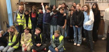 Wymiana uczniów między Gronau Comprehensive School a szkołą partnerską De Troubadour Zdjęcie przedstawia grupę około 25 uczennic w strefie wejściowej. Niektóre siedzą na podłodze z przodu, inne stoją blisko siebie za nimi. Kilka uczennic ma na sobie żółte kamizelki odblaskowe, inne kurtki lub bluzy z kapturem. Wiele z nich patrzy w stronę kamery, niektóre uśmiechają się lub machają. Na ścianie za grupą wisi duży, okrągły zegar, a pod nim znajdują się zamknięte drzwi. Po lewej stronie zdjęcia widać szatnię z mnóstwem kurtek i kilkoma parasolami, po prawej półki ze słuchawkami i wiszącą lampą. Pomieszczenie wygląda jak foyer muzeum lub wystawy.