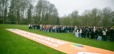 Um grande grupo de alunos está de pé numa área verde e forma uma longa fila. Em primeiro plano, encontra-se uma grande faixa cor de laranja com as palavras "Dignidade humana - juntos contra o racismo". As pessoas estão próximas umas das outras e, na sua maioria, vestem casacos. Ao fundo, vêem-se árvores e um céu nublado. A cena mostra uma grande concentração de alunos ao ar livre.