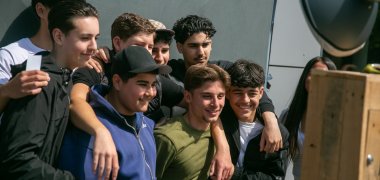 Vários alunos e alunas estão bem próximos uns dos outros em frente a uma cabine fotográfica e olham na sua direção. Alguns têm os braços à volta uns dos outros. Em primeiro plano, vê-se uma parte da cabine fotográfica; ao fundo, uma parede clara.