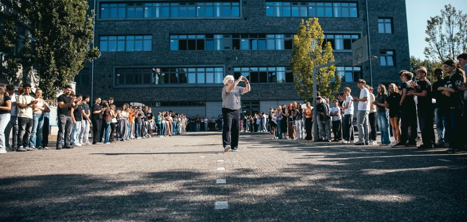 Rămas bun în fața comunității școlare, care a format un șir de oameni Ursula Steuer trece zâmbind pe lângă un lung șir de oameni care îi fac cinste în curtea școlii generale din Gronau. Elevii, profesorii și personalul școlii stau de ambele părți ale aleii, aplaudă și îi însoțesc în timpul intrării sale. În fundal se vede clădirea școlii sub un cer senin.