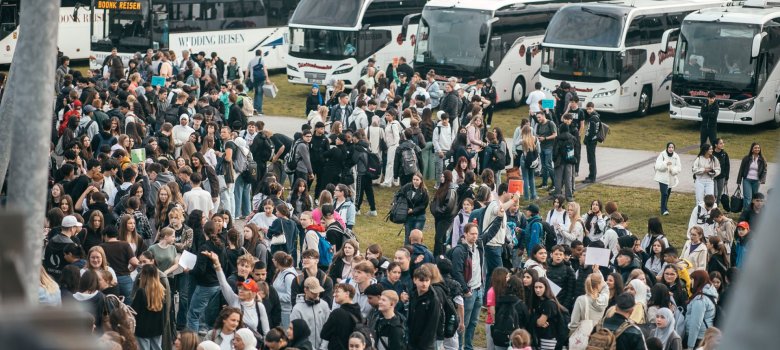 Vedere de sus asupra parcării de lângă Bürgerhalle: între 17 autocare de turism se adună sute de elevi și profesori de la Școala Generală din Gronau. Aceștia stau grupați, poartă rucsacuri, țin în mâini pancarte colorate cu numele grupurilor și se pregătesc pentru plecarea în comun spre Norderney.