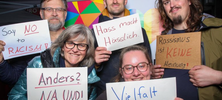 Päť ľudí rôzneho veku pózuje pred farebnou kulisou so srdcom v farbách dúhy. Do objektívu držia vlastnoručne vytvorené tabuľky s nápismi ako „Rozmanitosť spája“, „Rasizmus nie je názor“, „Nenávisť zohavuje“ a „Say NO to RACISM!“. Skupina stojí tesne vedľa seba a usmieva sa. Fotografia vznikla pri fotobúdke na festivale Európa a zachytáva ľudí rôznych generácií s rôznymi vyhláseniami proti rasizmu a vylúčeniu.