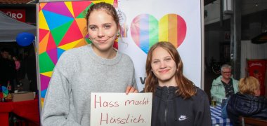 Dve mladé účastníčky stoja pred farebnou stienou s srdcom v farbách dúhy. Usmievajú sa do kamery a spoločne držia tabuľku s nápisom „Nenávisť robí človeka škaredým“. V pozadí sú vidieť miesta na sedenie a ďalší návštevníci a návštevníčky festivalu Európa.
