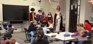 Učenci petega razreda sedijo za mizami v božično okrašeni učilnici, medtem ko Sinterklaas in njegovi pomočniki v pisanih kostumih stojijo pred učilnico. Otroci radovedno gledajo naprej in so navdušeni nad obiskom in majhnimi darili.