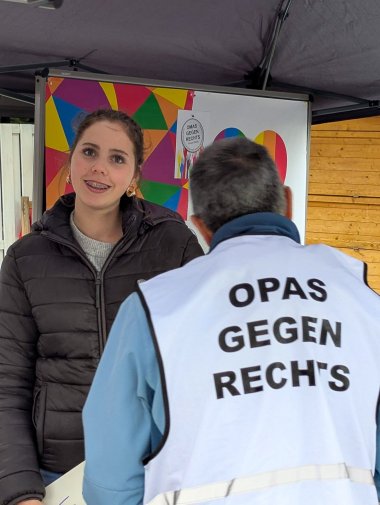 Bir çardak altında, bir kız öğrenci gökkuşağı renklerinde bir kalp desenli renkli bir panonun önünde duruyor. Ön planda, arkadan görünen ve üzerinde “Sağcı Karşıtı Büyükanneler” yazan açık renkli bir yelek giyen bir kişi var. İkisi birbirleriyle konuşuyor gibi görünüyor. Arka planda stantın diğer unsurları da görülüyor; bunların arasında girişimin bir afişi, ahşap bir kaplama ve çardak parçaları yer alıyor. Resim, Avrupa Festivali kapsamında nesiller arası bir sohbet anını gösteriyor.