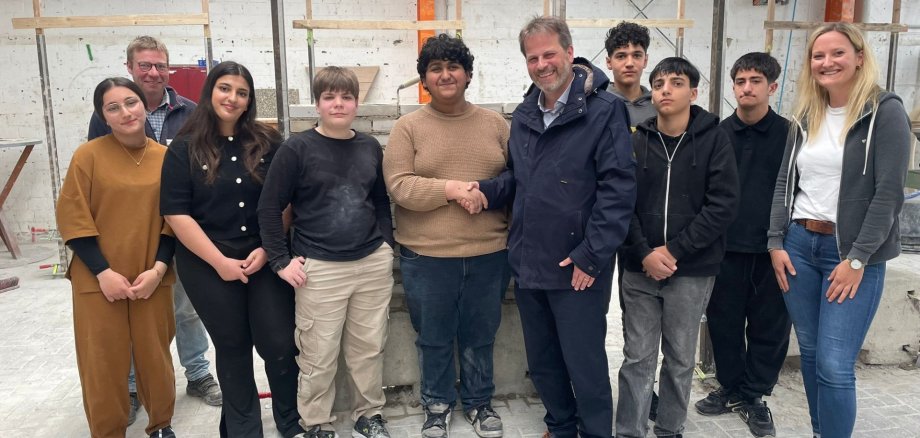 Dokuz genç ve üç yetişkinden oluşan bir grup, tuğla örme ve inşaat işlerinin yapıldığı bir atölyede duruyor. Arka planda tuğla duvarlar ve ahşap çerçeveler görülüyor. Ortada, koyu renk ceketli bir adam bej kazaklı bir öğrenciyle el sıkışıyor. Gençler gündelik kıyafetler giyiyor, bazılarının kıyafetlerinde ve ayakkabılarında pratik çalışmalardan kalma toz izleri görülüyor. Sarı saçlı bir kadın en sağda duruyor ve kameraya gülümsüyor. Grubun çoğu kameraya dostça ya da tarafsız bir şekilde bakıyor.