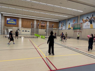 Mehrere Jugendliche spielen Badminton in einer großen Sporthalle. Die Halle ist mit verschiedenen Spielfeldern und Linien markiert. Einige Schülerinnen und Schüler stehen mit Schlägern auf dem Feld, während andere sich bewegen oder auf den Ballwechsel konzentrieren. An den Wänden hängen Sportbanner und Basketballkörbe.