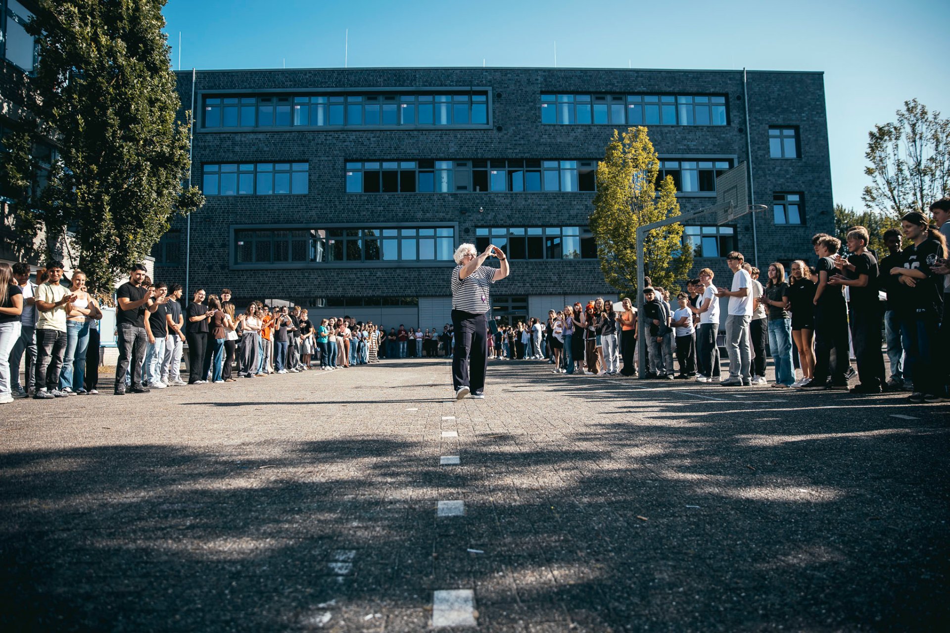 Ursula Steuer geht lächelnd durch ein langes Spalier auf dem Schulhof der Gesamtschule Gronau. Schülerinnen und Schüler, Lehrkräfte und Mitarbeitende stehen auf beiden Seiten des Weges, applaudieren und begleiten ihren Einzug. Im Hintergrund ist das Schulgebäude unter einem hellen Himmel zu sehen.